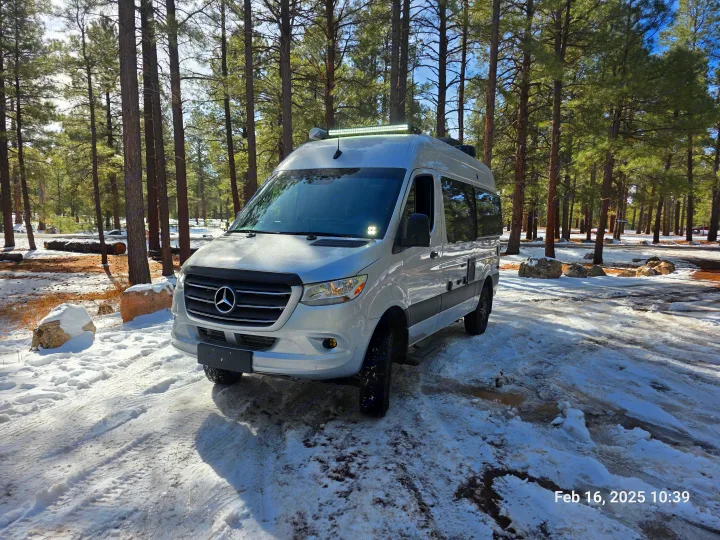 RV rental: A silver camper van parked in a snowy pine forest, surrounded by tall trees and white snow. Winter scene.. Thor Tranquility 19M 2025