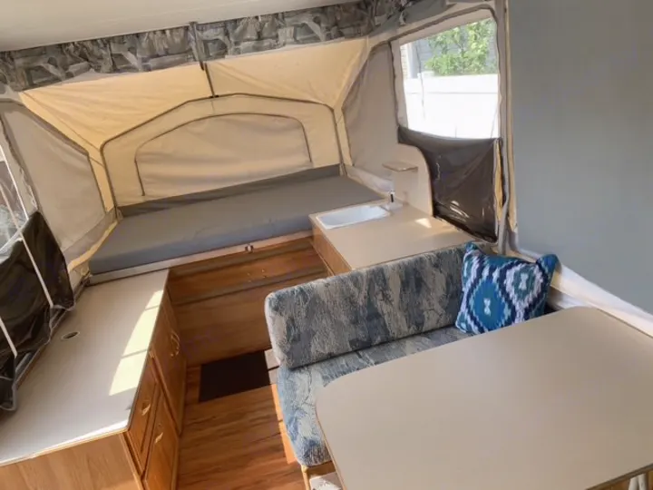 Inside an RV rental pop-up camper: view of the bed, compact kitchenette with sink, and a dinette with a patterned pillow.. Forest River Palomino 2000