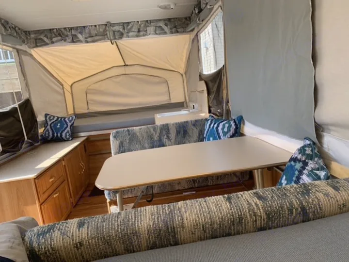 Pop-up RV rental interior: dining table, blue-patterned pillows, and beds, framed by canvas walls and wooden cabinets.. Forest River Palomino 2000