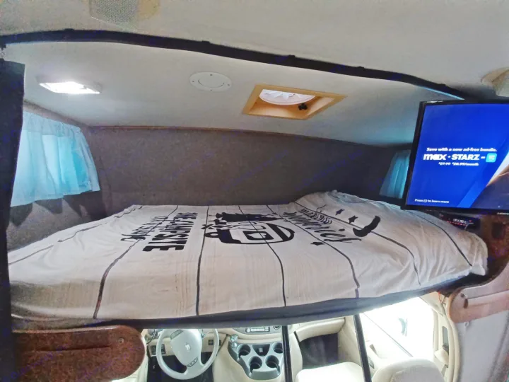 RV rental interior: cozy overhead bunk bed above the cab, featuring a TV, ceiling vent, and curtain.. Winnebago Outlook 2007