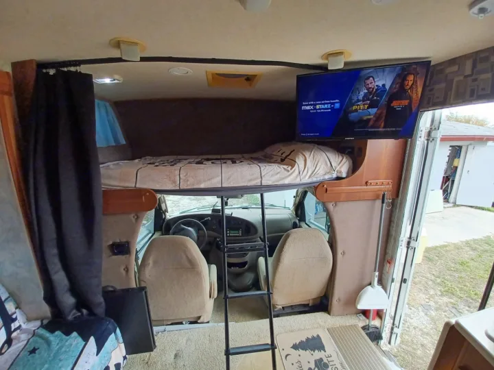 RV rental interior shows a cozy cab-over bunk bed above driver seats, with a TV, ladder, and cleaning tools.. Winnebago Outlook 2007