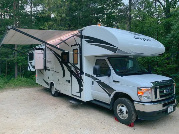 This white Jayco rv rental is set up for camping with its illuminated awning extended, parked on dirt in the woods.. Jayco Redhawk 2025