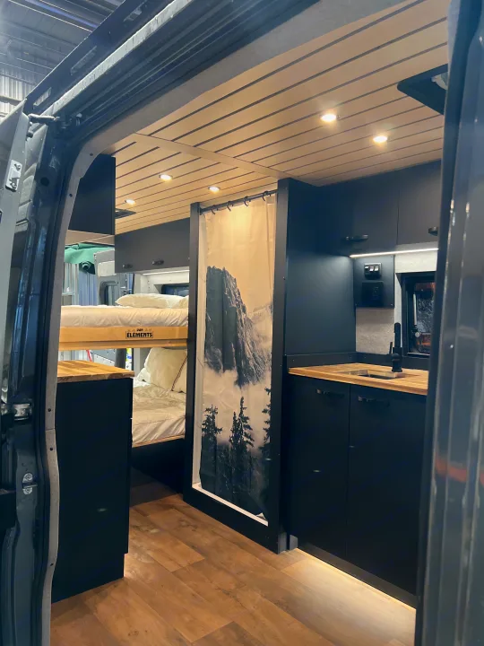Explore this modern RV rental: a dark-themed camper van interior with bunk beds, compact kitchen, and nature shower curtain.. Dodge Sportsman 2025