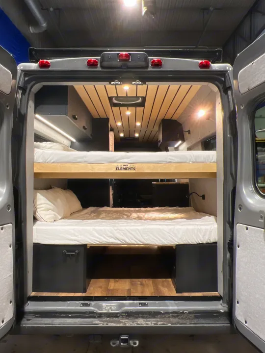 Inside a modern rv rental camper van: two cozy bunk beds with clean white bedding. Bright interior lighting & wood accents.. Dodge Sportsman 2025