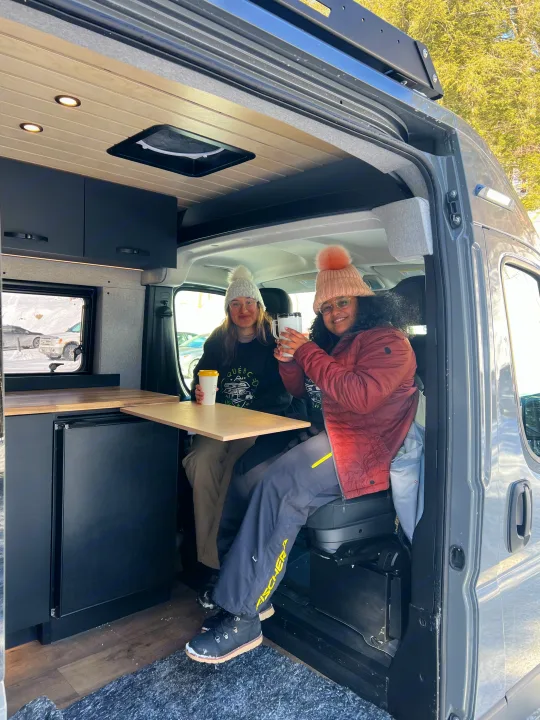 Two friends enjoy warm drinks inside a cozy RV rental, perfect for winter trips in Quebec's snowy landscapes.. Dodge Sportsman 2025