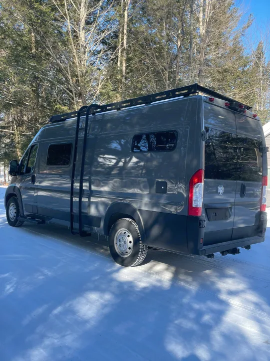 A modern gray RV rental camper van with a roof rack and ladder, parked in a sunny, snowy forest.. Dodge Sportsman 2025