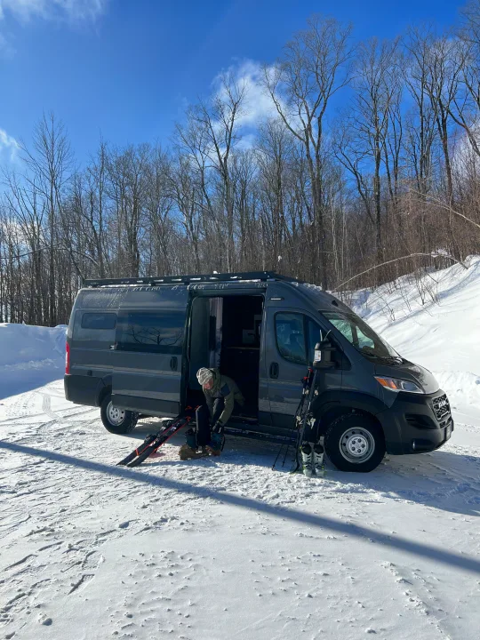 Getting ready for winter adventure! A person puts on ski boots beside a grey RV rental parked in deep snow.. Dodge Sportsman 2025