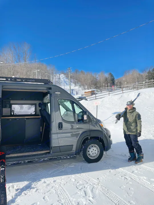 A skier with skis stands next to a custom grey RV rental van, parked at a snowy mountain ski resort.. Dodge Sportsman 2025