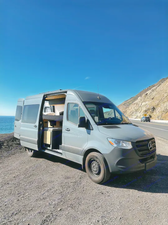 Grey RV rental camper van by ocean on a coastal highway, side door open showing interior. Ideal for scenic travel & van life.. Mercedes-Benz Sprinter 2024