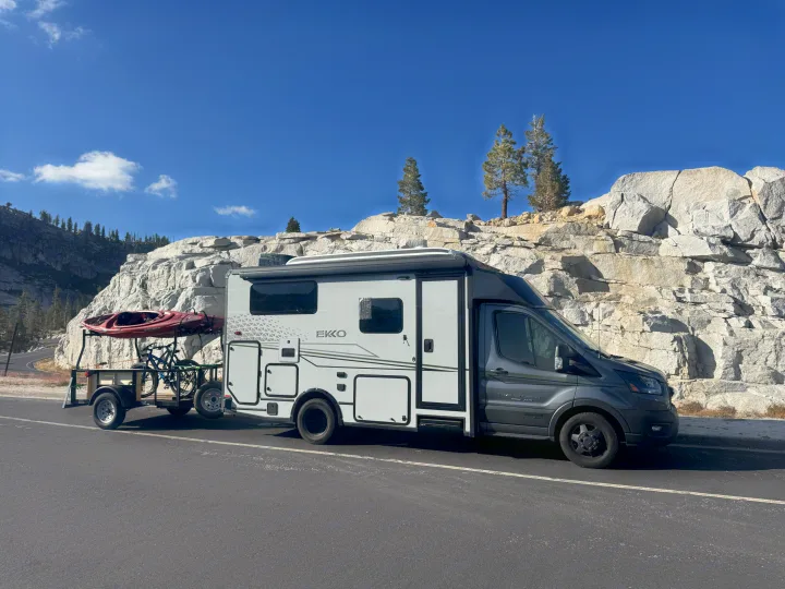 Grey Winnebago Ekko **rv rental** with a trailer carrying kayaks & bikes, parked on a scenic mountain road beside rocky cliffs.. Winnebago Ekko 2023