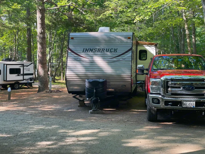 A white Innsbruck travel trailer and a red Ford truck are parked at a shaded, dirt campsite. This RV rental offers a perfect nature getaway.. Gulf Stream Innsbruck 2014