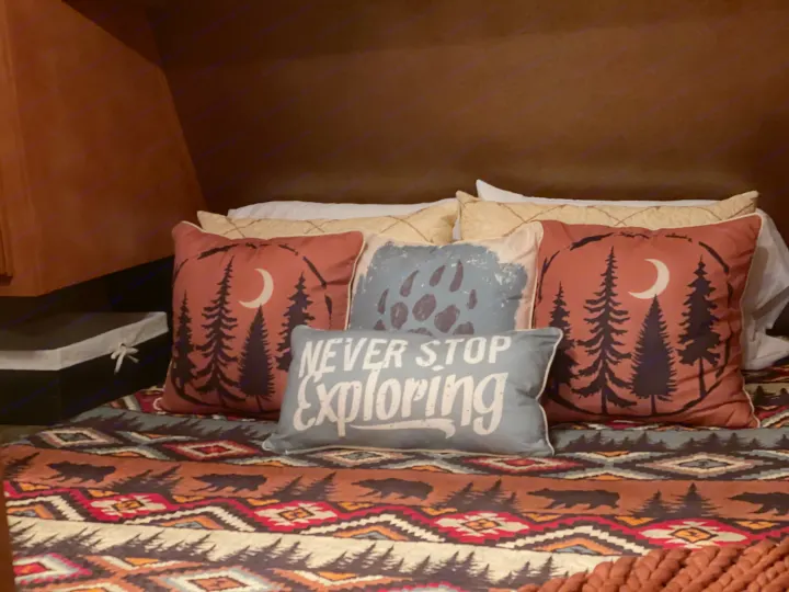 Cozy RV rental bed featuring moon & pine tree pillows, plus a "NEVER STOP EXPLORING" pillow. Rustic bear & tree quilt. Ideal for travel comfort.. Gulf Stream Innsbruck 2014
