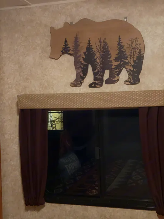 Cozy RV rental interior displays a large wooden bear wall decoration with etched forest trees above a window with brown curtains.. Gulf Stream Innsbruck 2014