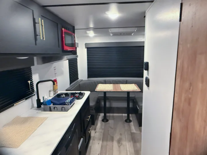 Compact RV rental interior featuring a modern kitchen with dark cabinets, a red microwave, and a cozy dinette area with black blinds.. Forest River Wildwood FSX 2022
