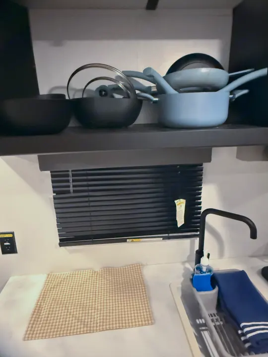 An RV rental kitchen with stacked pots and pans, a neat sink area, blinds, and a checkered dishtowel on the counter.. Forest River Wildwood FSX 2022