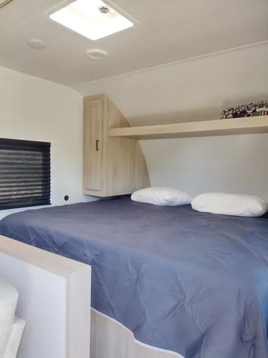 Cozy RV rental bedroom with a dark blue bed, pillows, overhead storage, and a window for comfortable travel.. East to West Della Terra LE 2024
