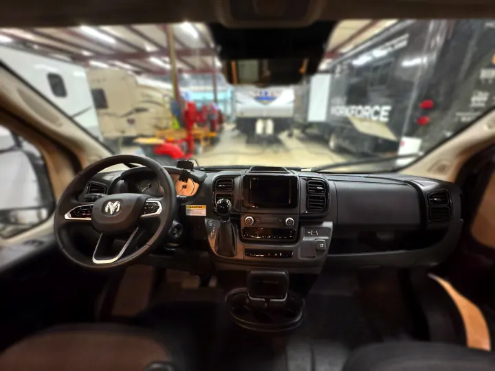 View from inside a Ram van: dashboard, steering wheel. Blurry background shows RVs and trucks in a warehouse. Ideal for rv rental.. Winnebago Solis 2021