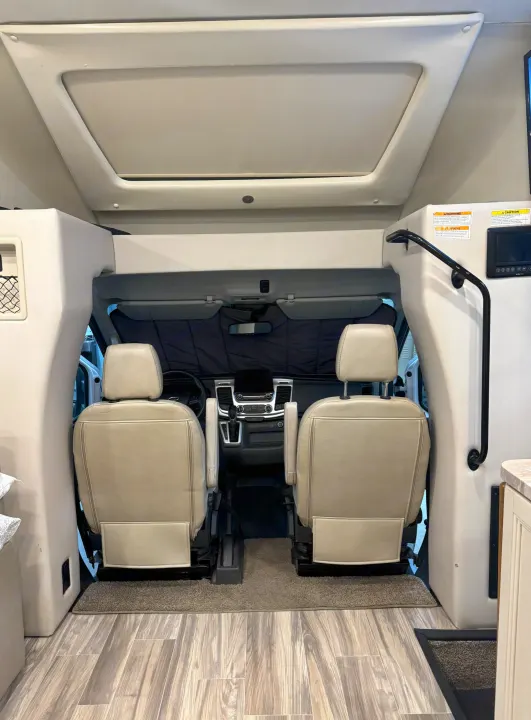 Interior view of an RV rental, showing the cockpit with two tan seats, dashboard, and an overhead bunk/storage area.. Winnebago Solis 2021
