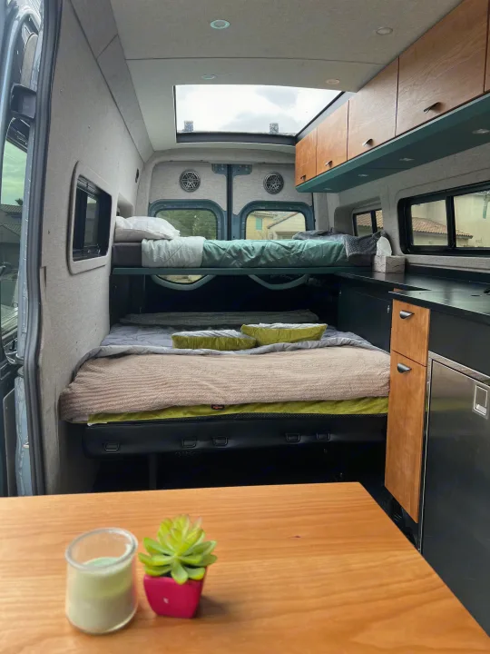 RV rental interior view: bunk beds, lower bed, compact kitchen with fridge/cabinets, and bright skylight above.. Off Highway Van Off Highway Van Class B 2024
