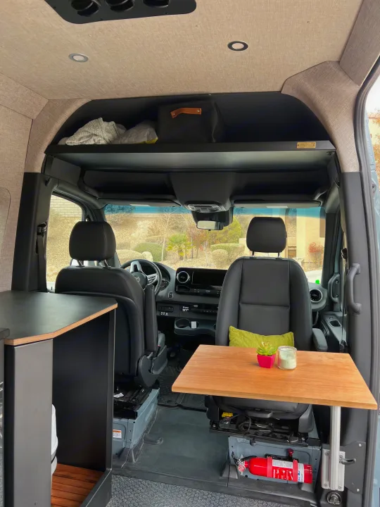 This RV rental camper van interior has an overhead shelf, wood table, and safety features like a fire extinguisher.. Off Highway Van Off Highway Van Class B 2024
