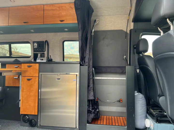 Explore this RV rental's compact interior: a full kitchen with wooden cabinets, fridge, and a clever shower area.. Off Highway Van Off Highway Van Class B 2024