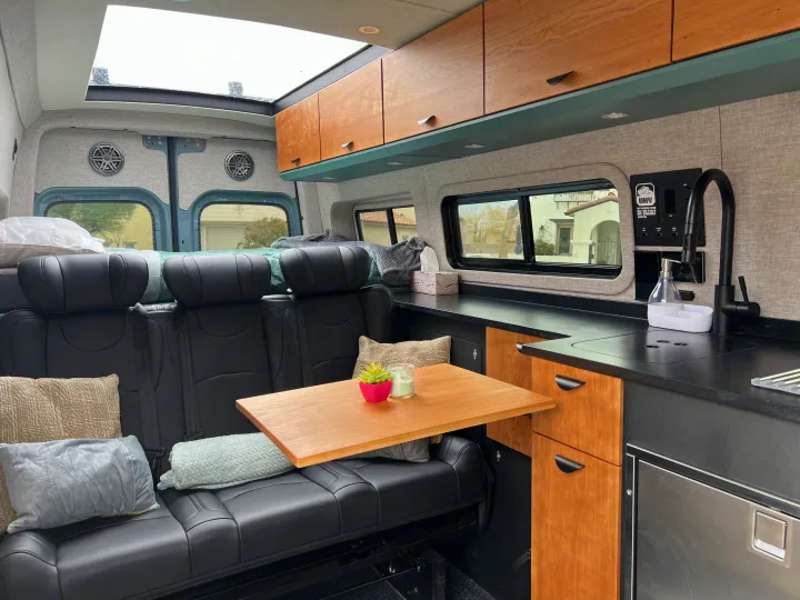Spacious RV rental interior featuring black leather seating, a wooden table, and a well-equipped kitchen for trips.. Off Highway Van Off Highway Van Class B 2024