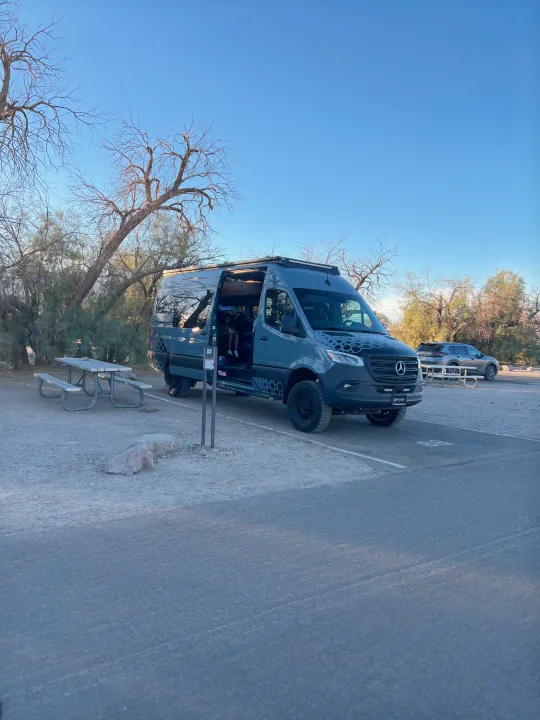 Grey Mercedes Sprinter camper van at campsite 88 with an open door, ready for an RV rental trip. A picnic table is nearby.. Off Highway Van Off Highway Van Class B 2024