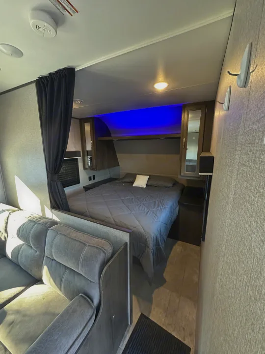 RV rental interior: A bedroom with a dark gray bed & blue LED lights, separated by a curtain from the living area with a gray sofa.. Jayco Jay Flight SLX 2020