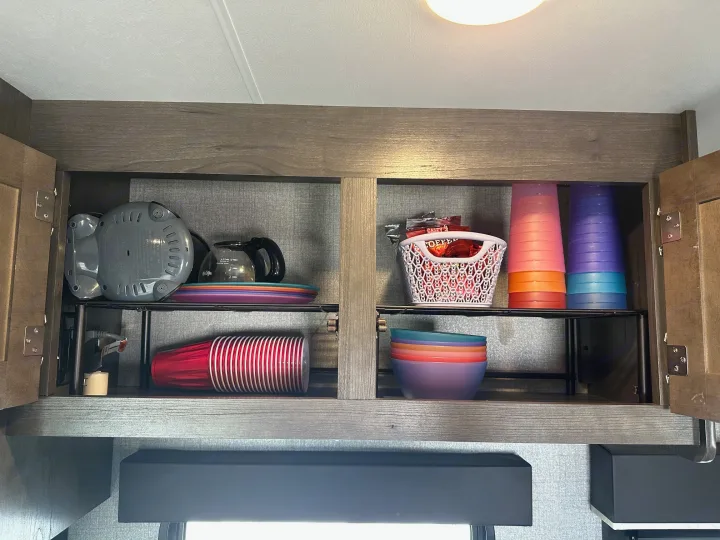 An open RV rental cabinet displaying an organized kitchen setup with a coffee maker, stacked plates, colorful cups, bowls, and snacks.. Jayco Jay Flight SLX 2020