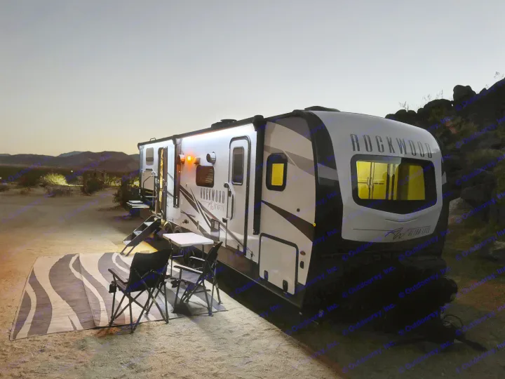 Enjoying a peaceful night in the desert with a cozy rv rental setup. Lights illuminate the travel trailer, rug, and chairs.. Forest River Rockwood Ultra Lite 2022