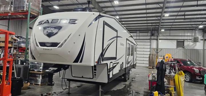 A white Sabre Cobalt fifth-wheel RV is inside a well-lit service bay, potentially for rv rental preparation or maintenance.. Forest River Sabre 2019