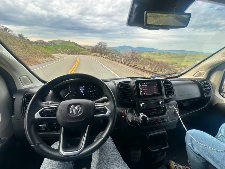 Driving an RV rental van through scenic green hills on a winding road, dashboard and passenger visible under a cloudy sky.. Winnebago Solis 2023
