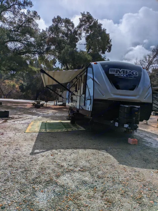 An MPG Ultra-Lite RV rental is parked at a gravel campsite with its awning extended and a green rug outside. Light precipitation visible.. Cruiser RV MPG Ultra Lite 2019