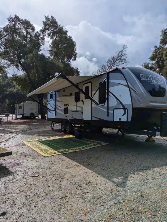 Spacious RV rental set up at a gravel campsite with an extended awning, green rug, trees, and cloudy sky.. Cruiser RV MPG Ultra Lite 2019