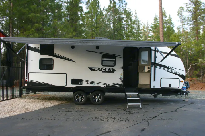 White & black Prime Time Tracer travel trailer RV rental parked on gravel, awning extended, steps down, amidst pine trees.. Forest River Tracer 2022