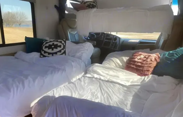 Spacious RV rental interior with multiple comfy beds, white sheets, and an overhead bunk. Scenic lake view outside.. Coachmen Pursuit 2021