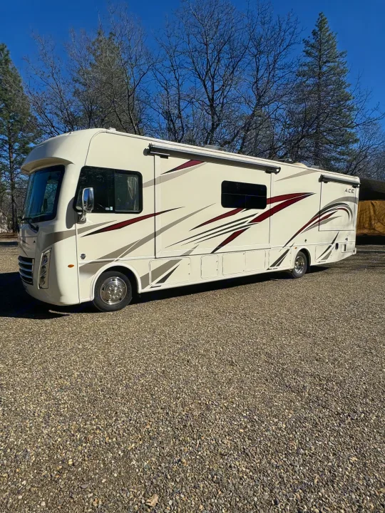 Cream-colored RV rental with maroon and gray stripes parked on a sunny gravel lot, surrounded by trees.. Thor A.C.E. 2019