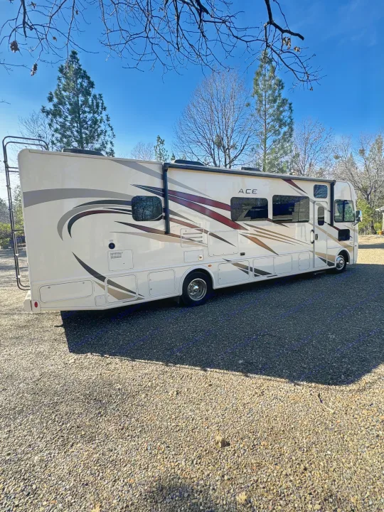 A tan ACE rv rental motorhome is parked on a sunny gravel driveway. The RV has decorative stripes, visible windows & ladder.. Thor A.C.E. 2019