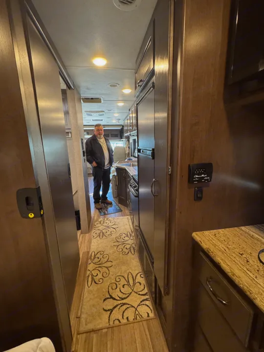 A man stands in the spacious interior of an RV rental, featuring a kitchen, cabinets, and warm lighting.. Thor A.C.E. 2019