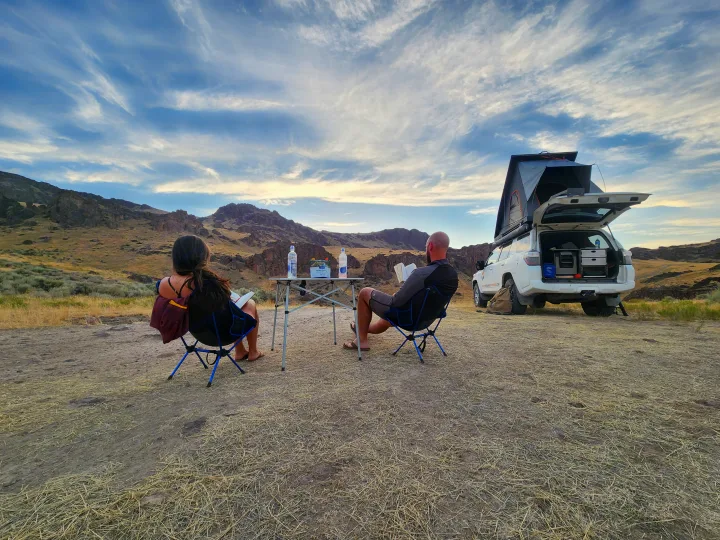 Couple unwinds outdoors, reading beside their white SUV RV rental with a rooftop tent, amidst a rugged landscape.. Toyota 4Runner 2021