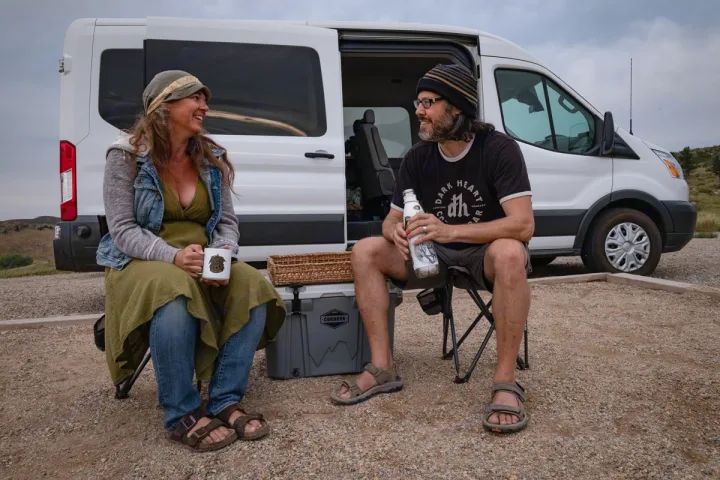 Smiling couple chats, relaxing in chairs next to their white RV rental van at a gravel campsite.. Ford Transit Custom 2015