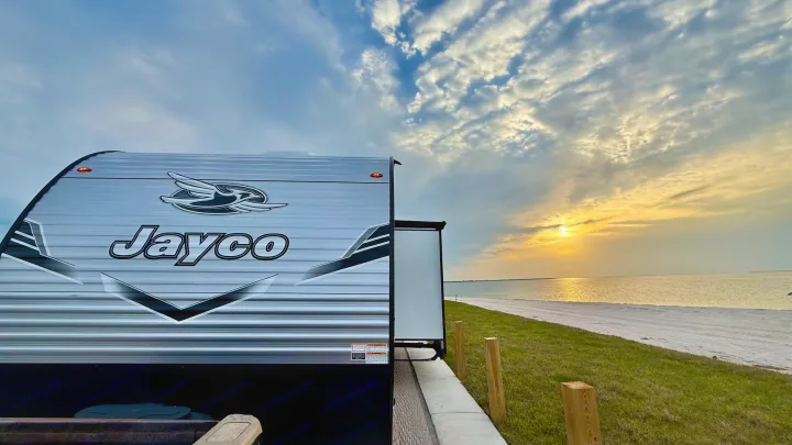 Experience tranquil evenings with an RV rental. A modern Jayco camper overlooks a serene beach and a beautiful sunset over the water.. Jayco 265BHS 2025