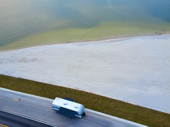 An RV rental is parked on a road, overlooking a wide sandy beach and calm bay waters, as seen from above.. Jayco 265BHS 2025