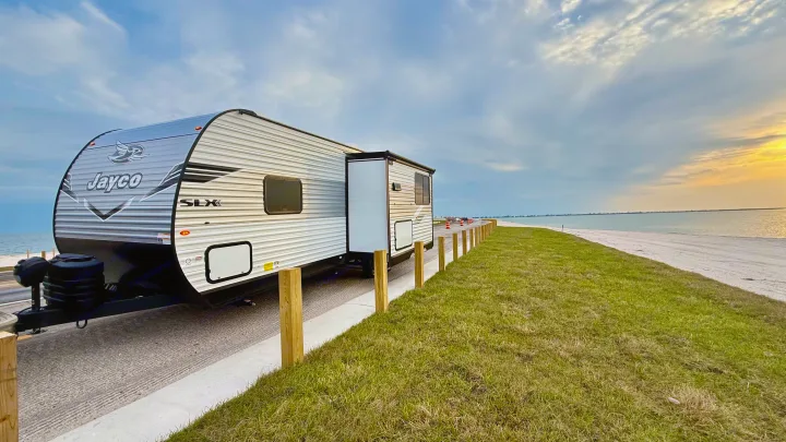 RV rental: Jayco SLX travel trailer by a grassy slope, sandy beach, and calm water under a cloudy sunset sky.. Jayco 265BHS 2025