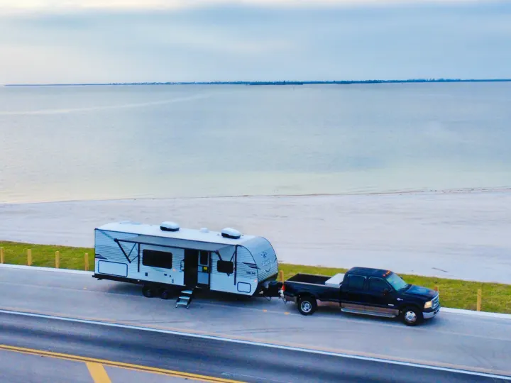 Scenic RV rental: A black pickup truck tows a white travel trailer, parked roadside with a sandy beach view and calm bay.. Jayco 265BHS 2025