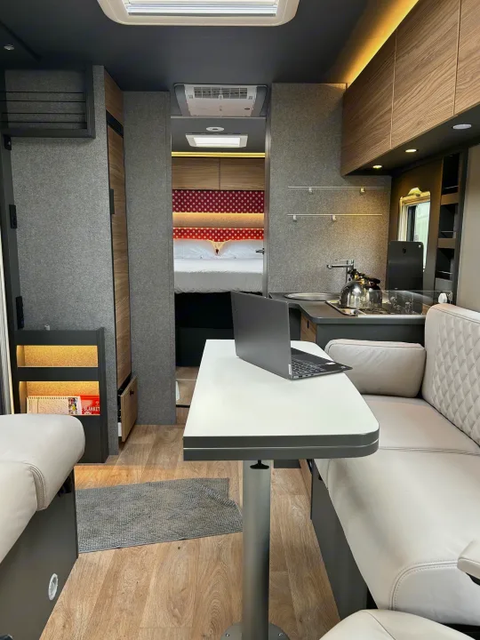 Modern rv rental interior showing a laptop on the table, a compact kitchen, and a cozy bedroom.. GlamperRV DBL 2026