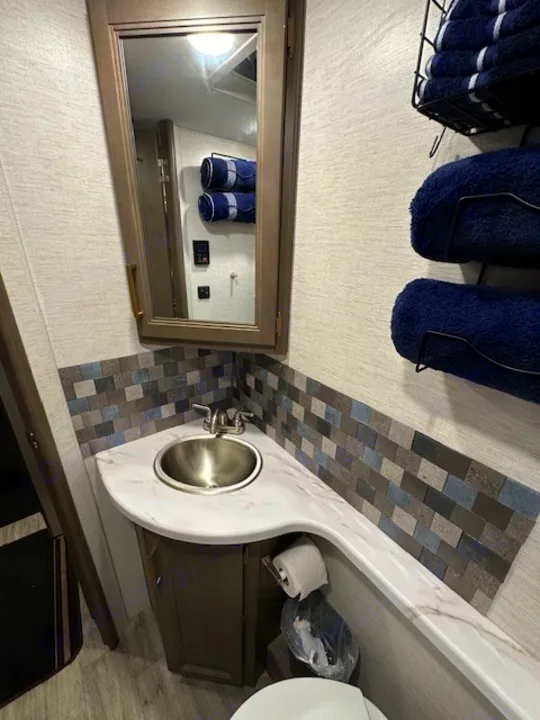 Compact RV rental bathroom featuring a corner sink, mirror, mosaic tile backsplash, and blue towels.. Thor Motor Coach Four Winds 22B 2025