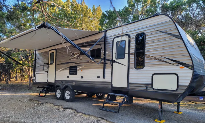 An RV rental travel trailer with an extended awning, parked in a peaceful wooded setting, ideal for camping.. Jayco Jay Flight 2018