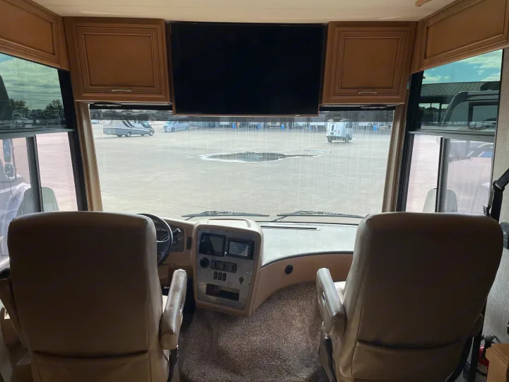RV rental interior: Cockpit view with dashboard, seats, overhead TV, and large windshield looking out at a wet lot.. Canyon Star Canyon Star Motorhome 2020