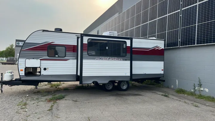 Modern RV rental, a travel trailer with extended slide, parked by a building covered in solar panels.. Prime Time Avenger 2024
